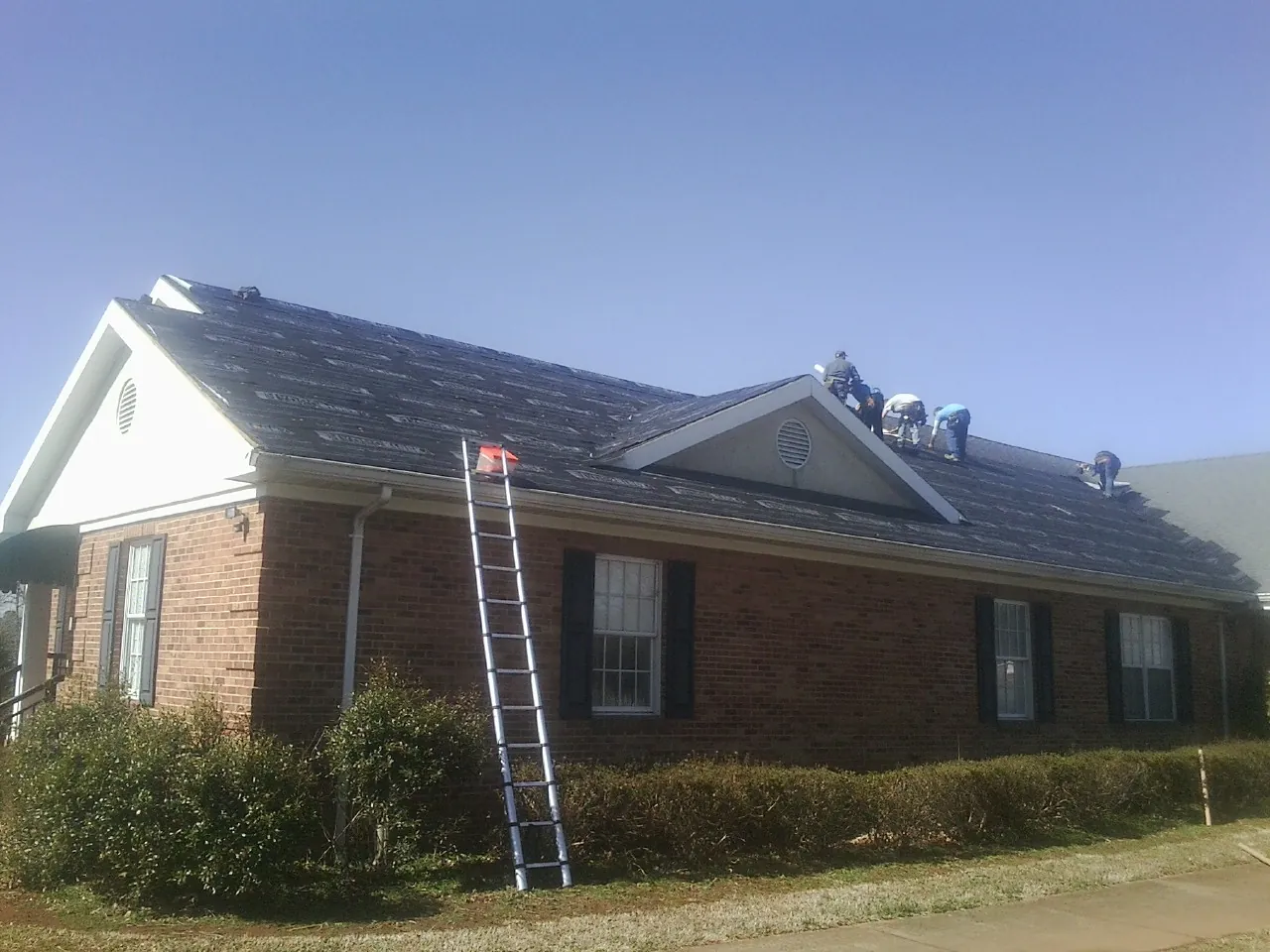 Crew tearing shingles off a brick ranch roof with a ladder set — Emergency Roof Repair work by our crew in Mira Loma