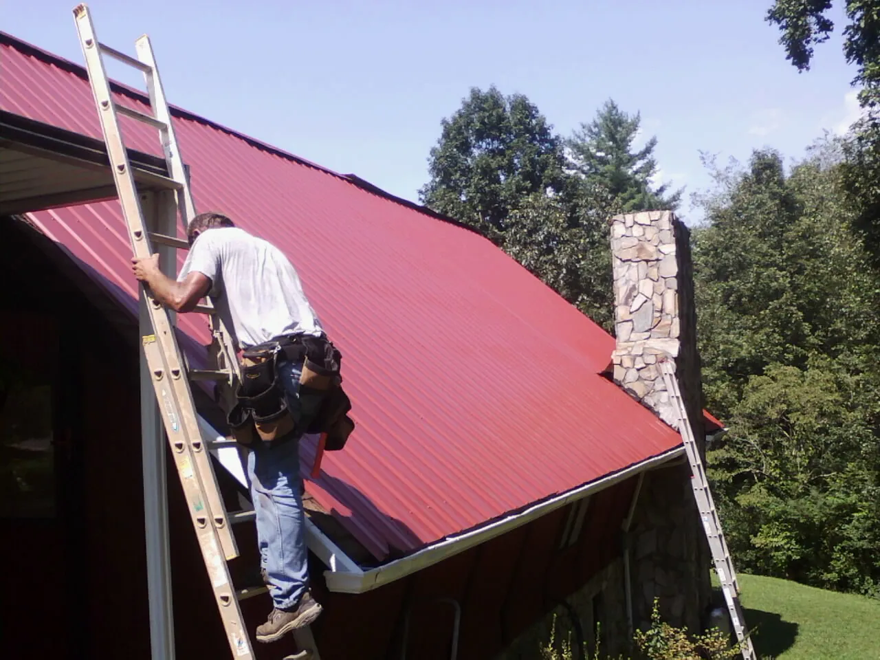 Roofer on a ladder beside a red metal roof and stone chimney — Soffit & Fascia Repair by Roofing Brothers in Mira Loma